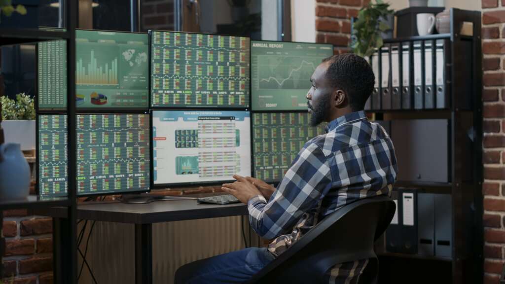 A Men Sitting on chair in from of many Computer Displays And Monitoring themm